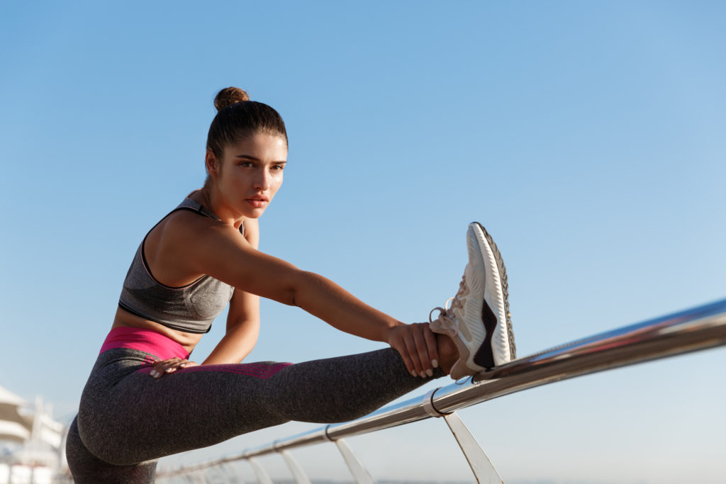 Image of attractive fit fitness girl workout on a pier. Sportswoman stretching legs and warming-up before jogging training on the seaside promenade.